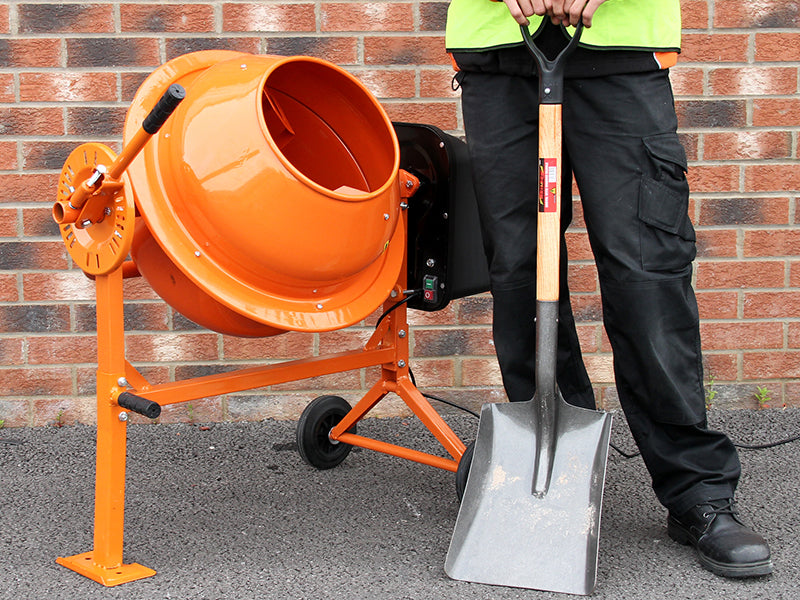 Malaxeur à béton orange vif sur un support, avec une pelle grise à côté, fond de briques rouges et sol en asphalte texturé