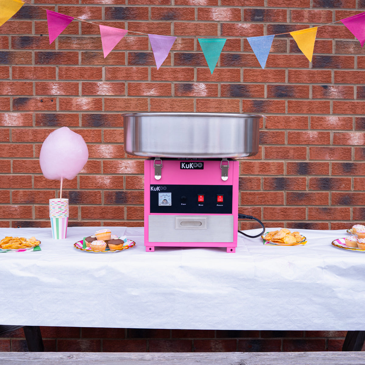 Machine à barbe à papa rose avec cuve en acier inoxydable, décorée de guirlandes colorées, sur une table avec des douceurs festives