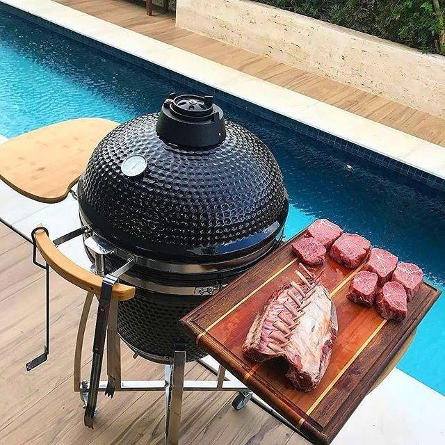 Barbecue au charbon de bois Kamado noir, 53,3 cm, avec viande grillée sur une planche en bois près d'une piscine bleue