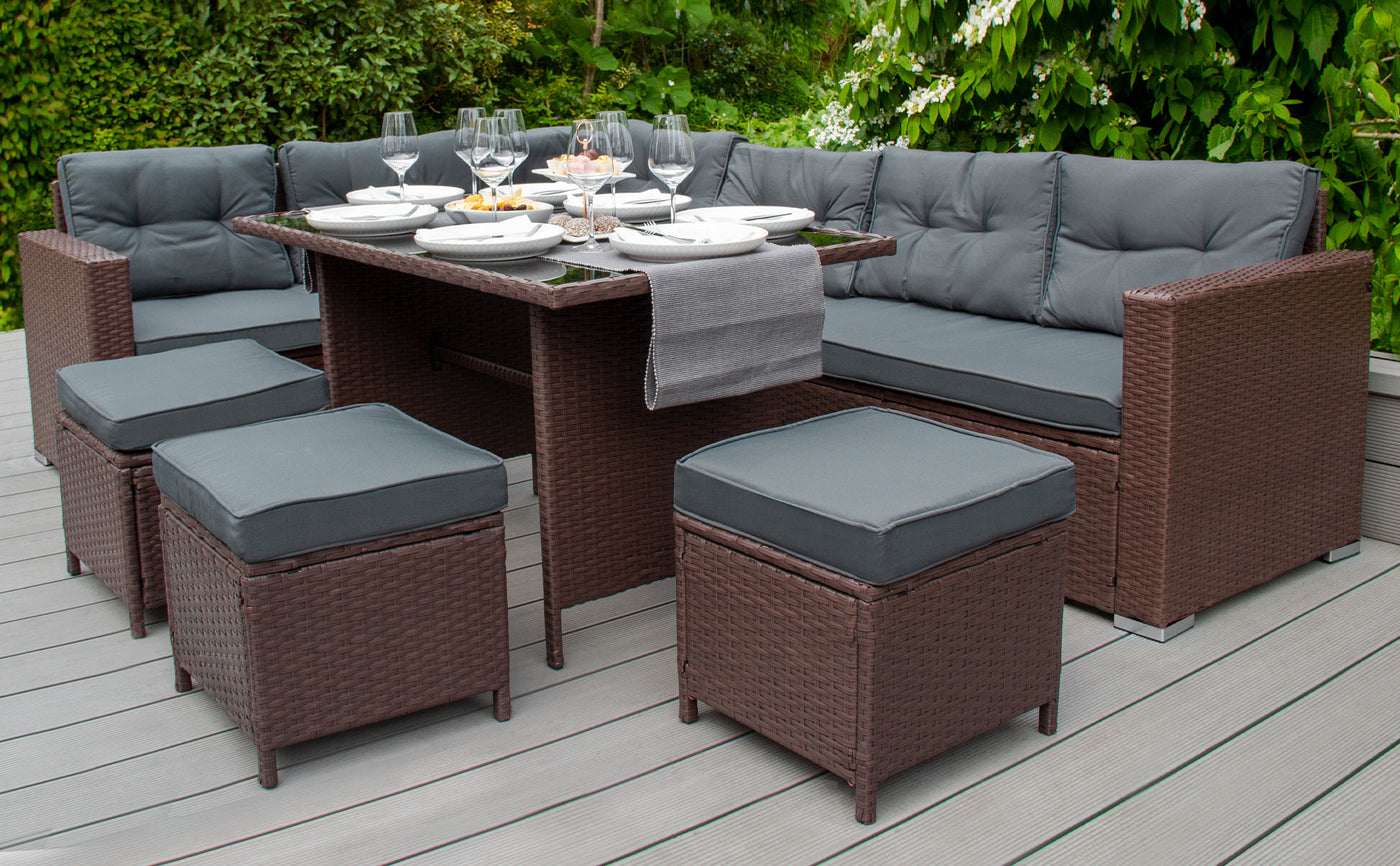 Salon de jardin en rotin marron avec coussins gris, table centrale, assiettes et verres, entouré de verdure luxuriante