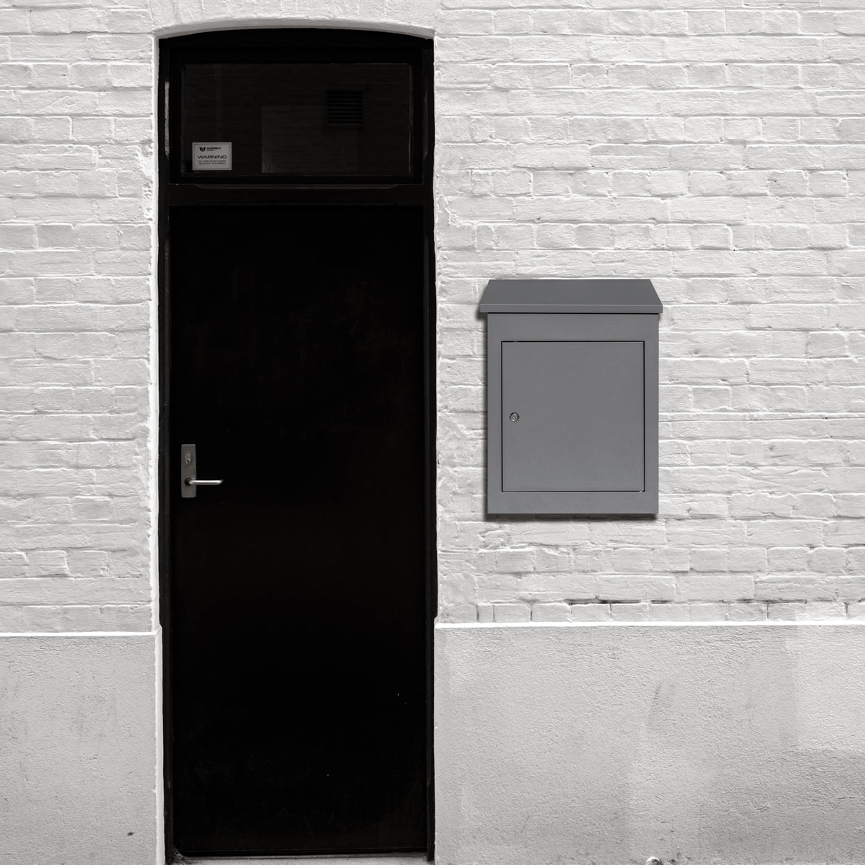 Boîte à colis anti-vol grise, design moderne et épuré, fixée à un mur blanc, à côté d'une porte noire élégante