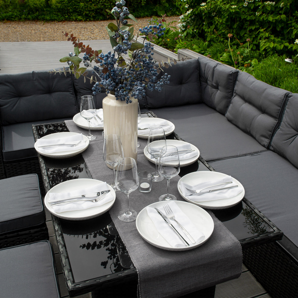Salon de jardin en rotin noir pour 9 personnes, avec coussins gris, table en verre, assiettes blanches et vase décoratif avec fleurs