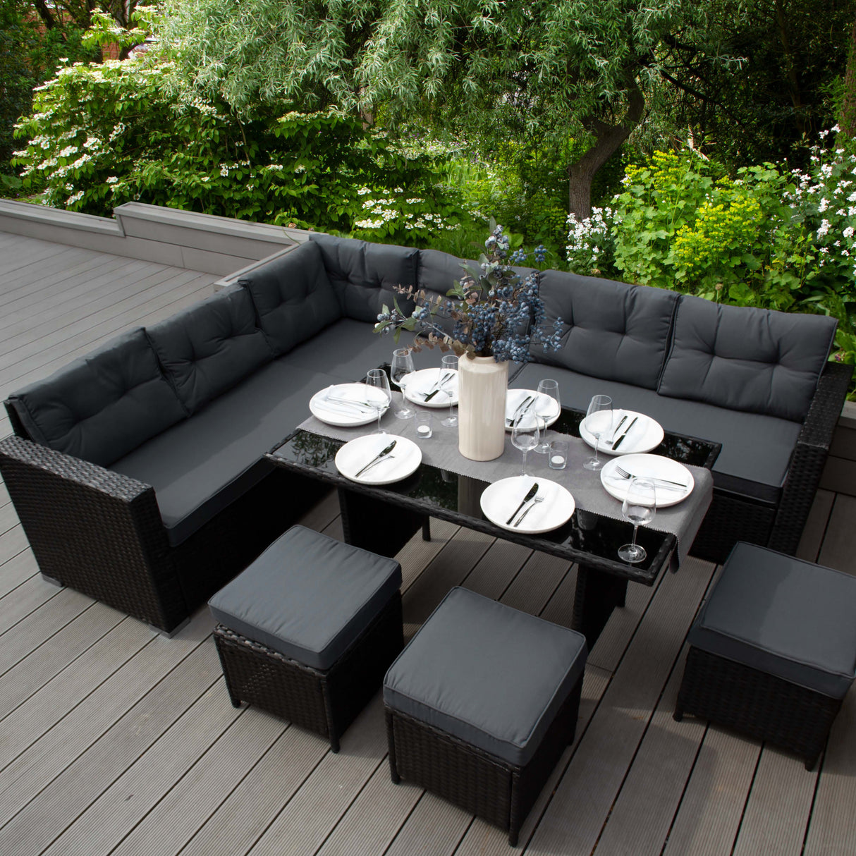 Salon de jardin en rotin noir pour 9 personnes, avec coussins gris, table en verre, et décor floral sur une terrasse en bois