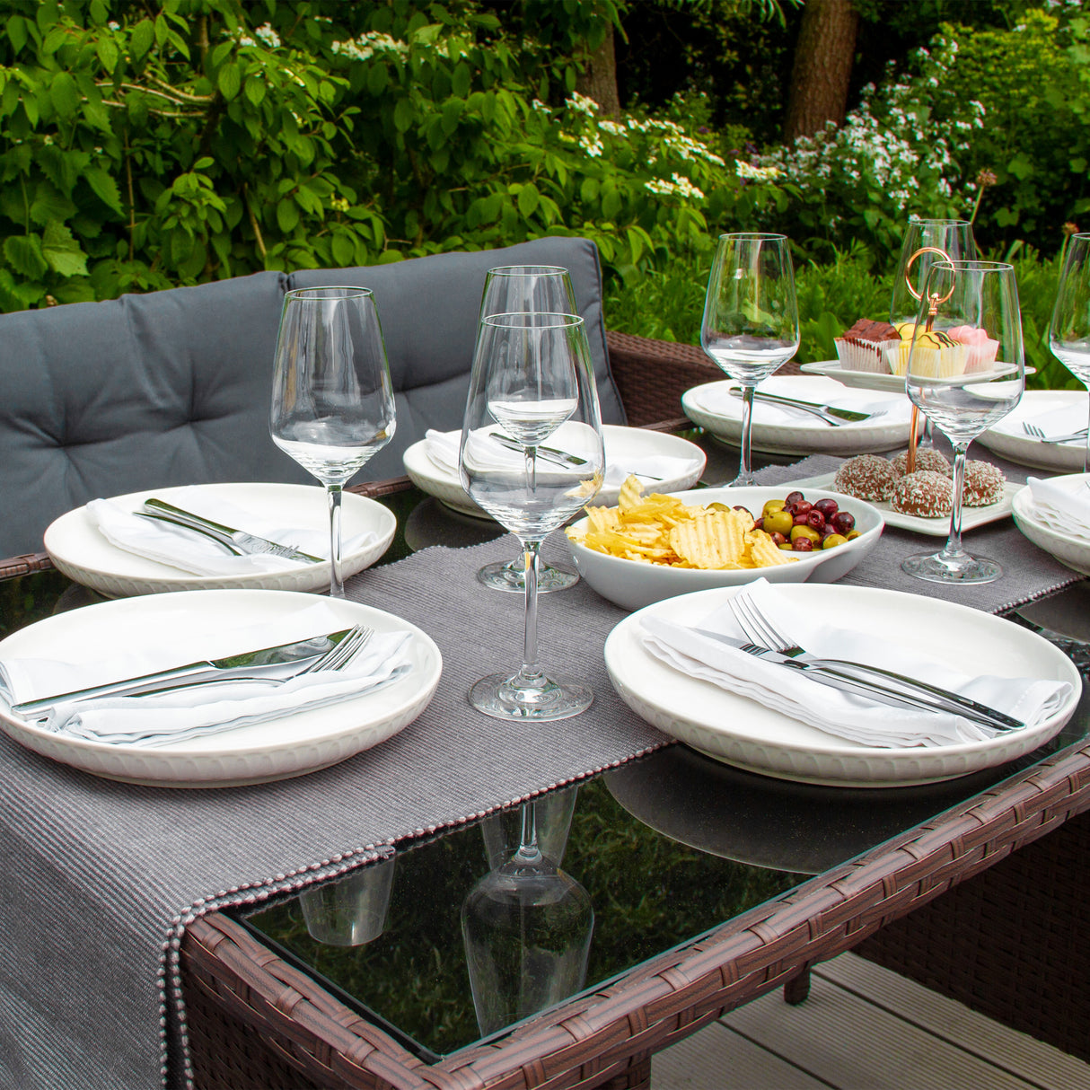 Salon de jardin en rotin marron pour 9 personnes, table en verre, assiettes blanches, couverts en acier, décor floral en arrière-plan