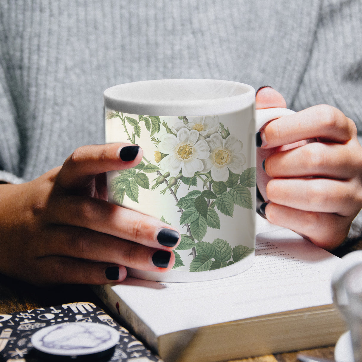Tasse blanche ornée de fleurs blanches et de feuillages verts, tenue par des mains avec des ongles noirs, sur une table en bois