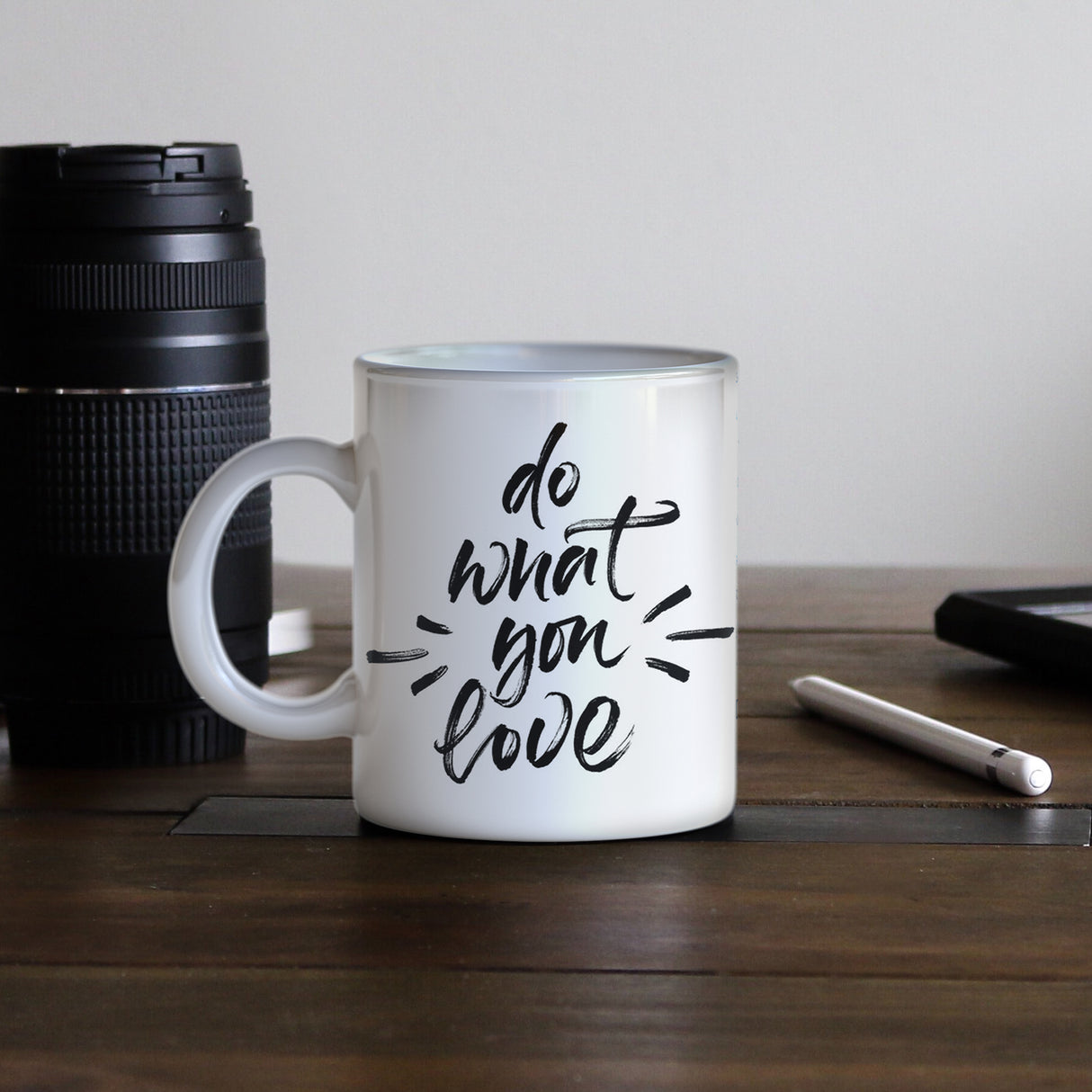 Tasse blanche en céramique avec inscription noire "do what you love", posée sur un bureau en bois avec un appareil photo et un stylo