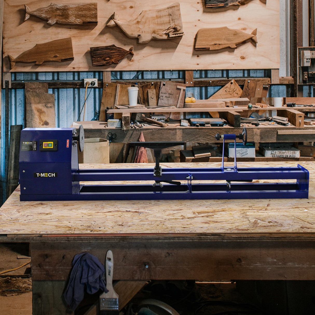 Tour à bois T-Mech en métal bleu, avec mandrin et accessoires, posé sur un établi en bois, entouré de chutes de bois