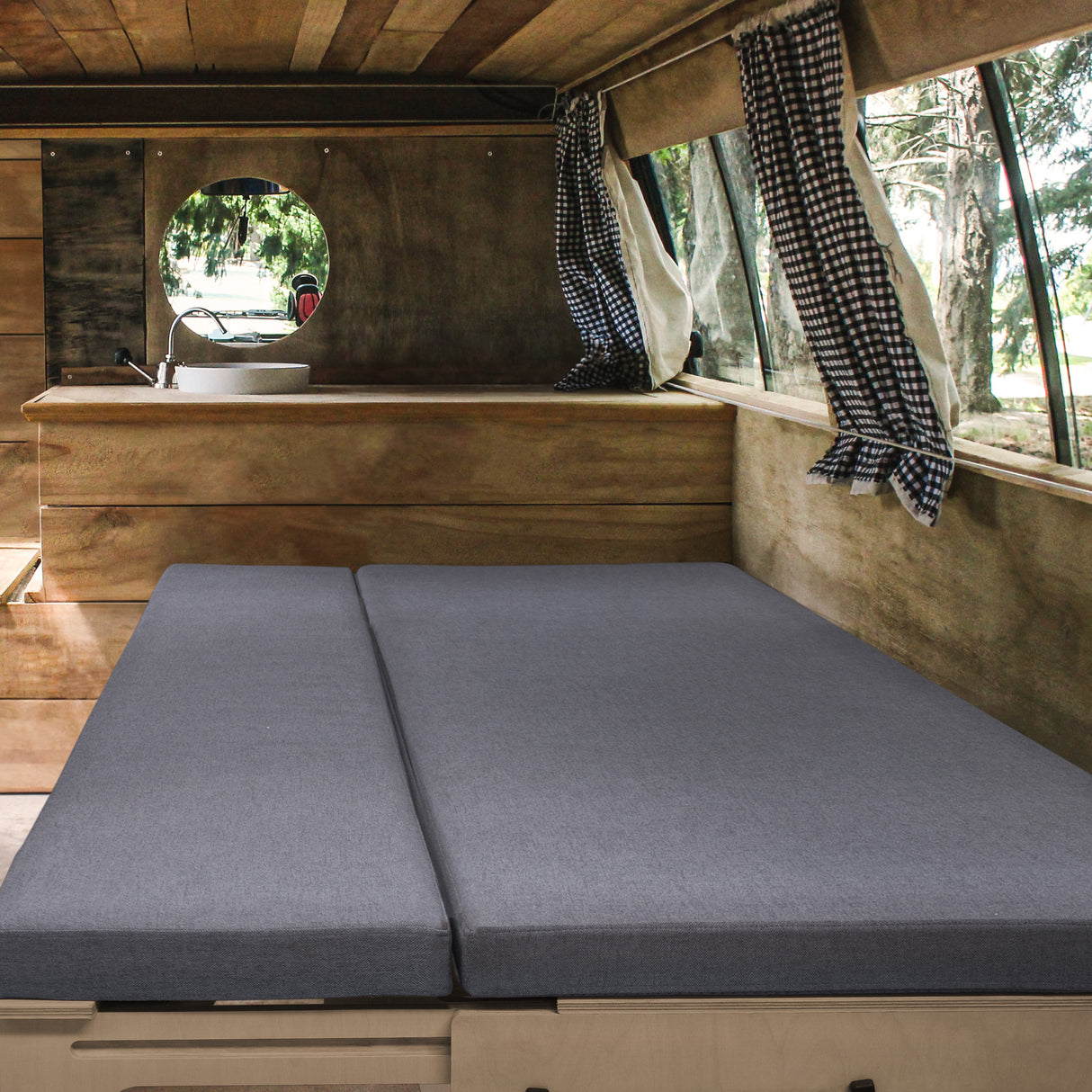 Canapé-lit gris en tissu doux, installé dans un camping-car en bois avec des rideaux à carreaux et une fenêtre ronde lumineuse