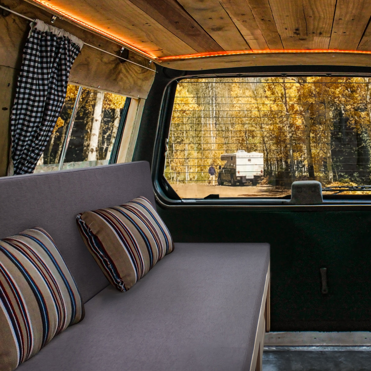 Canapé-lit gris avec coussins rayés, installé dans un camping-car, offrant une vue sur une forêt automnale à l'extérieur