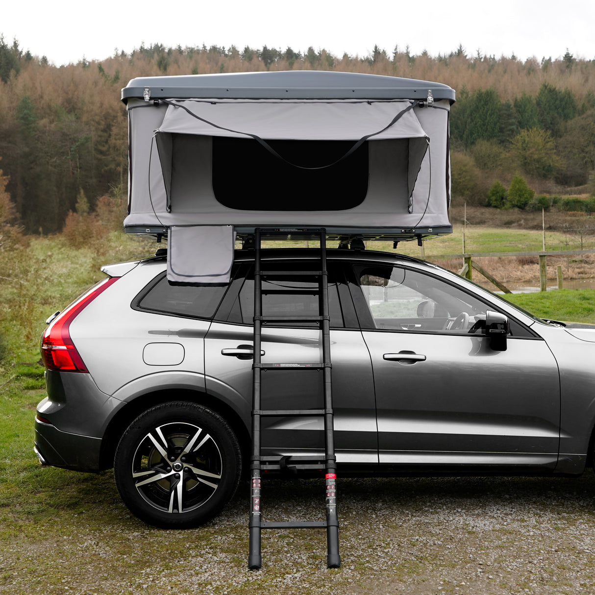 Tente de toit grise pour voiture avec échelle, installée sur un véhicule, entourée d'une nature verdoyante et d'arbres en arrière-plan