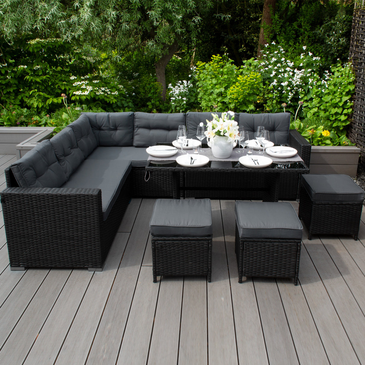 Salon de jardin en rotin noir pour 9 personnes, avec coussins gris, table en verre et poufs assortis, entouré de verdure luxuriante