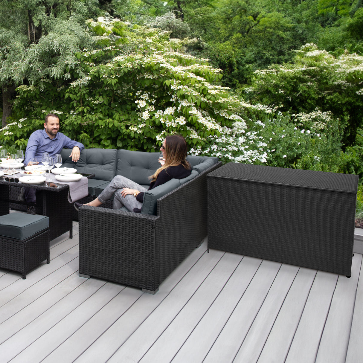 Salon de jardin en rotin noir pour 9 personnes, avec un coffre de rangement, installé sur une terrasse en bois, entouré de verdure