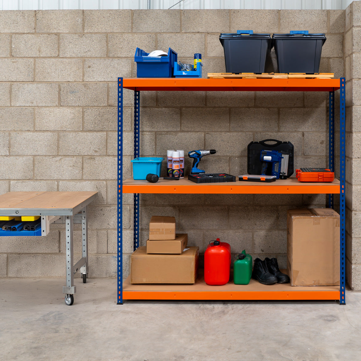 Rayonnage S-Rax en acier orange et bleu avec trois étagères, rempli de boîtes, outils, et cartons sur un mur en béton gris
