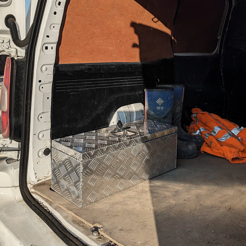 Caisse à outils en aluminium argenté, 35 L, avec motif en relief, placée dans le coffre d'un véhicule utilitaire, à côté de vêtements de travail