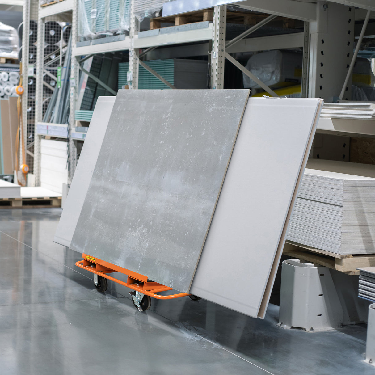 Chariot orange pour plaques de plâtre et tapis en caoutchouc, transportant des panneaux gris, sur un sol en béton dans un entrepôt bien organisé