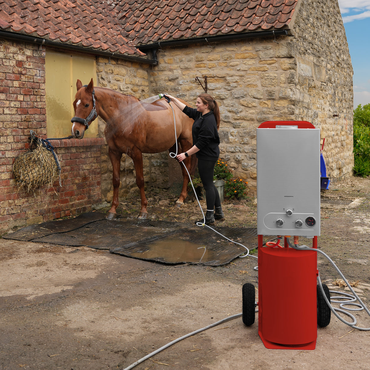 Douche de cheval portable deluxe rouge, avec un design moderne, utilisée pour laver un cheval dans une cour en pierre, sol humide et herbeux