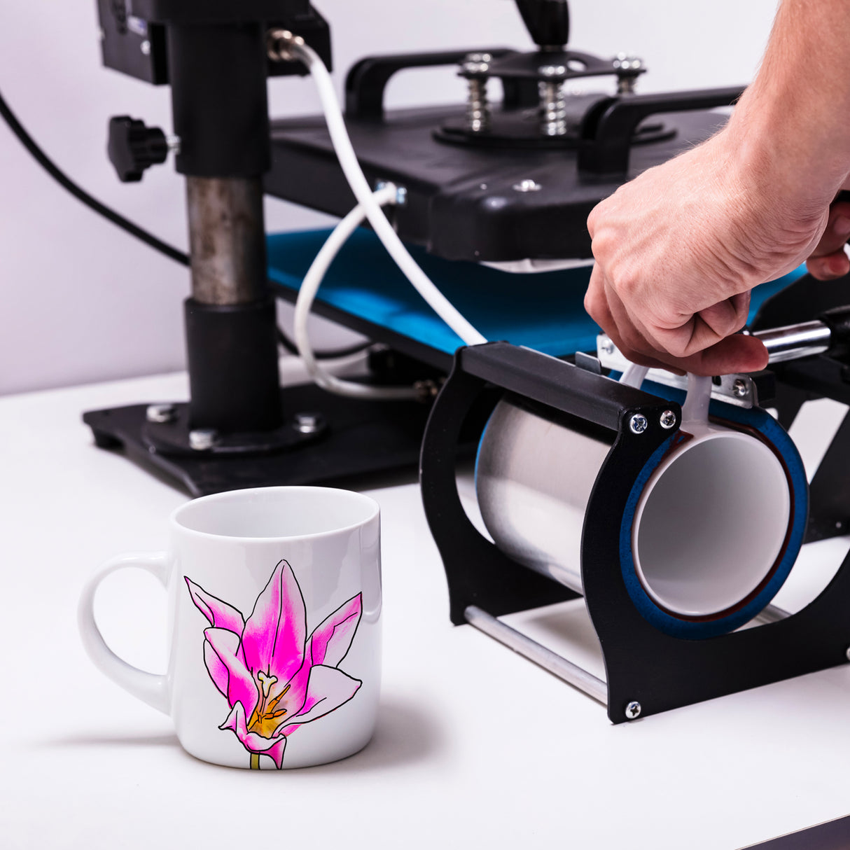 Tasse blanche avec un motif de fleur rose, à côté d'une machine de sublimation et d'un ruban thermorésistant sur une table blanche