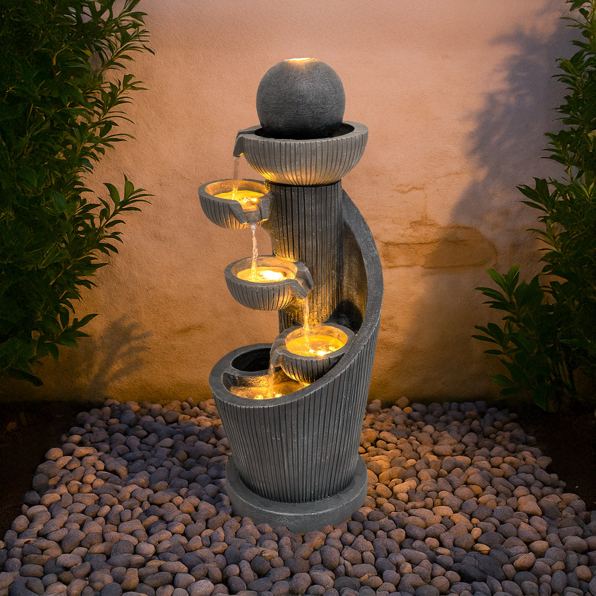 Fontaine de jardin en pierre grise avec éclairage LED, présentant des cascades en spirale et des sphères décoratives, entourée de galets