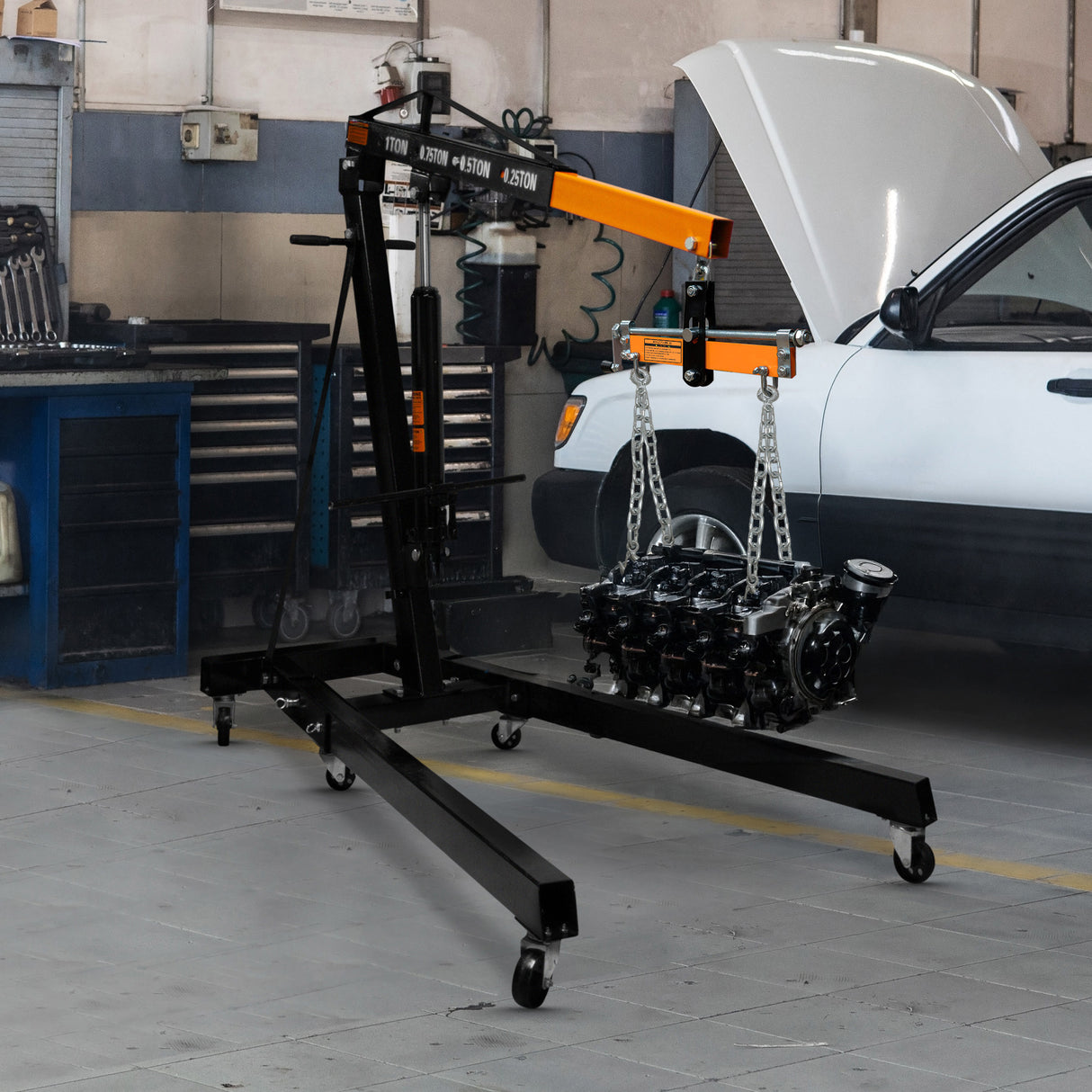 Grue de moteur noire avec bras orange, soulevant un moteur automobile, dans un garage avec outils et véhicule en arrière-plan