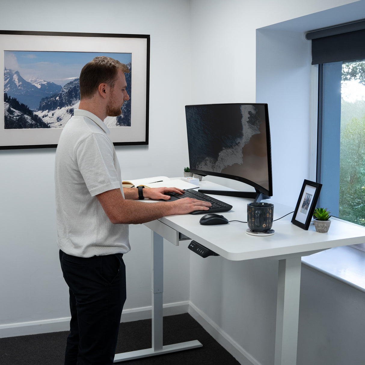 Bureau assis-debout électrique blanc avec tiroir, homme debout utilisant un ordinateur, décor moderne et lumineux, vue sur la nature