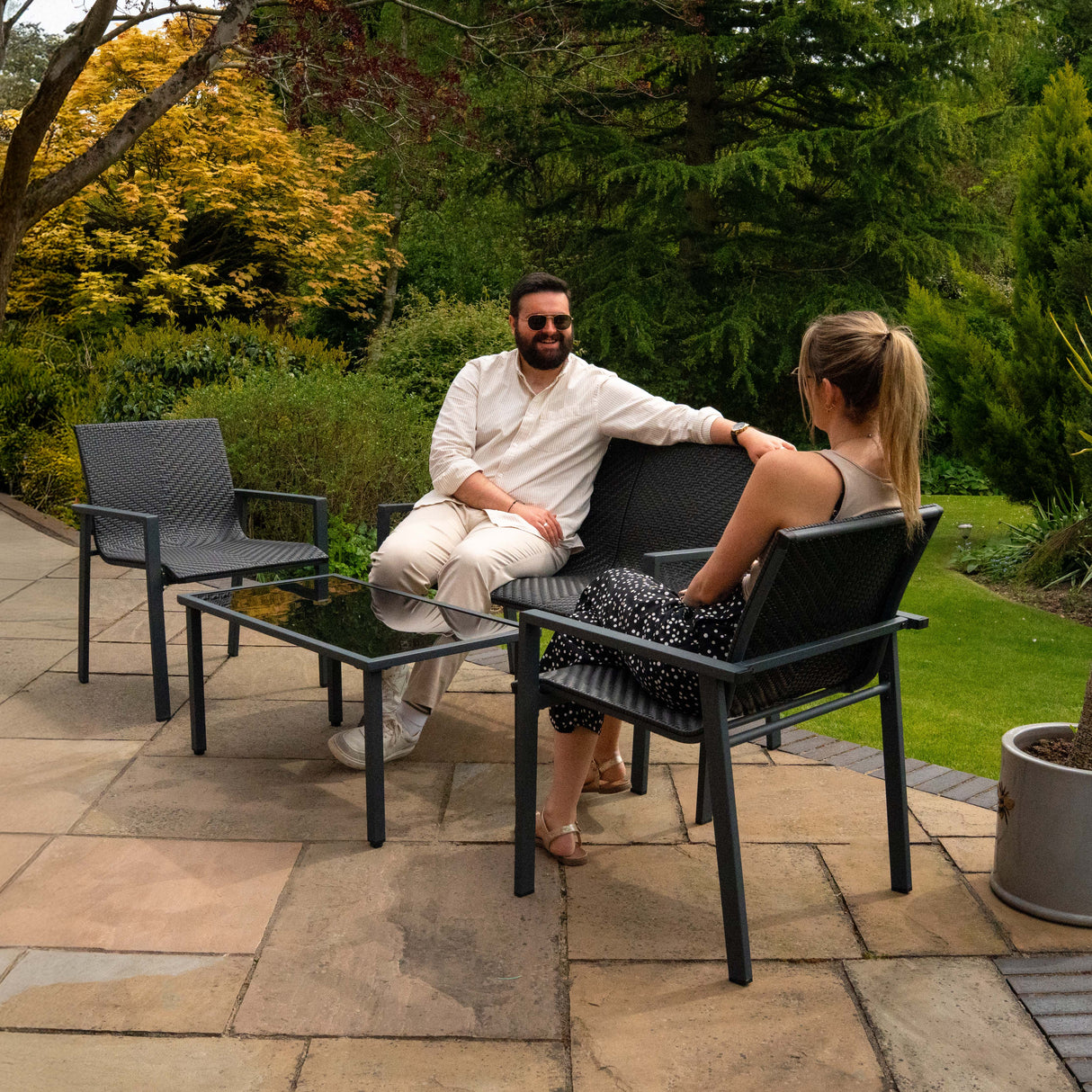 Ensemble de salon de terrasse en rotin noir, comprenant deux chaises et une table basse en verre, dans un jardin verdoyant