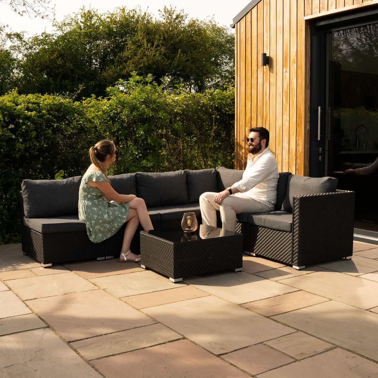 Salon de jardin modulaire en rotin noir avec coussins gris, composé de sept pièces, installé sur une terrasse en pierre naturelle