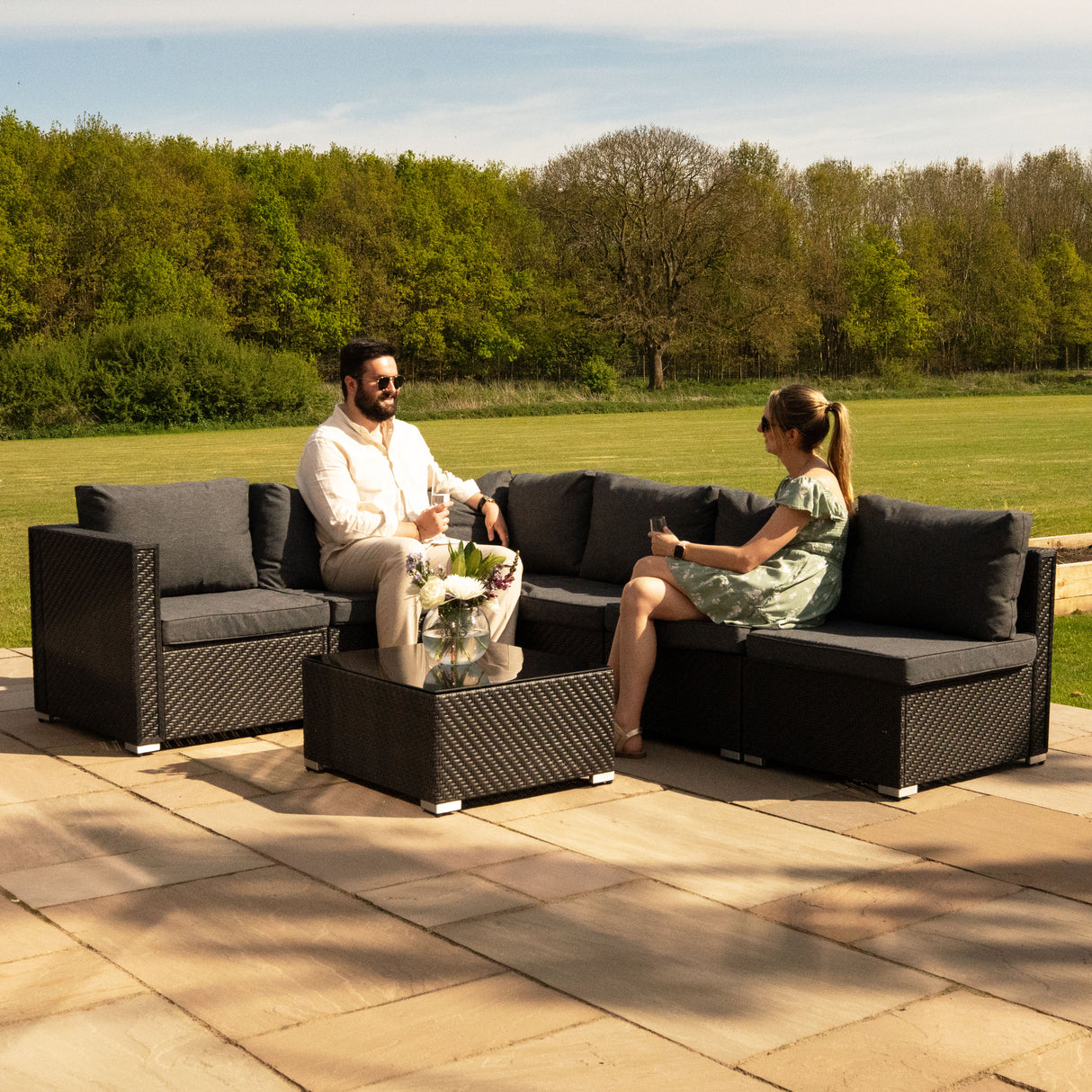 Salon de jardin modulaire en rotin noir, 7 pièces, avec coussins gris, installé sur une terrasse en pierre, entouré de verdure
