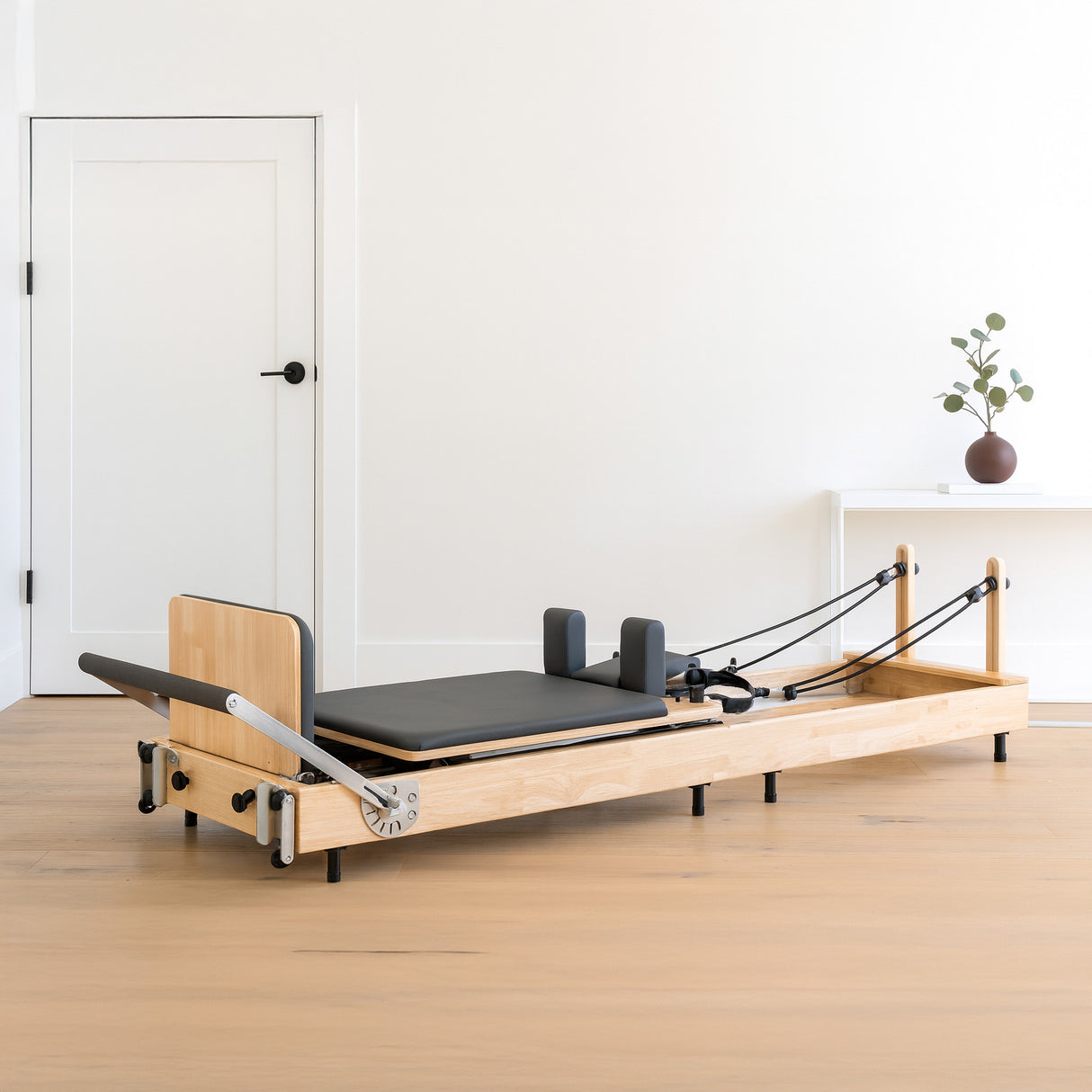 Machine reformer Pilates pliable en chêne clair, avec un coussin noir, des ressorts et des poignées, sur un sol en bois clair