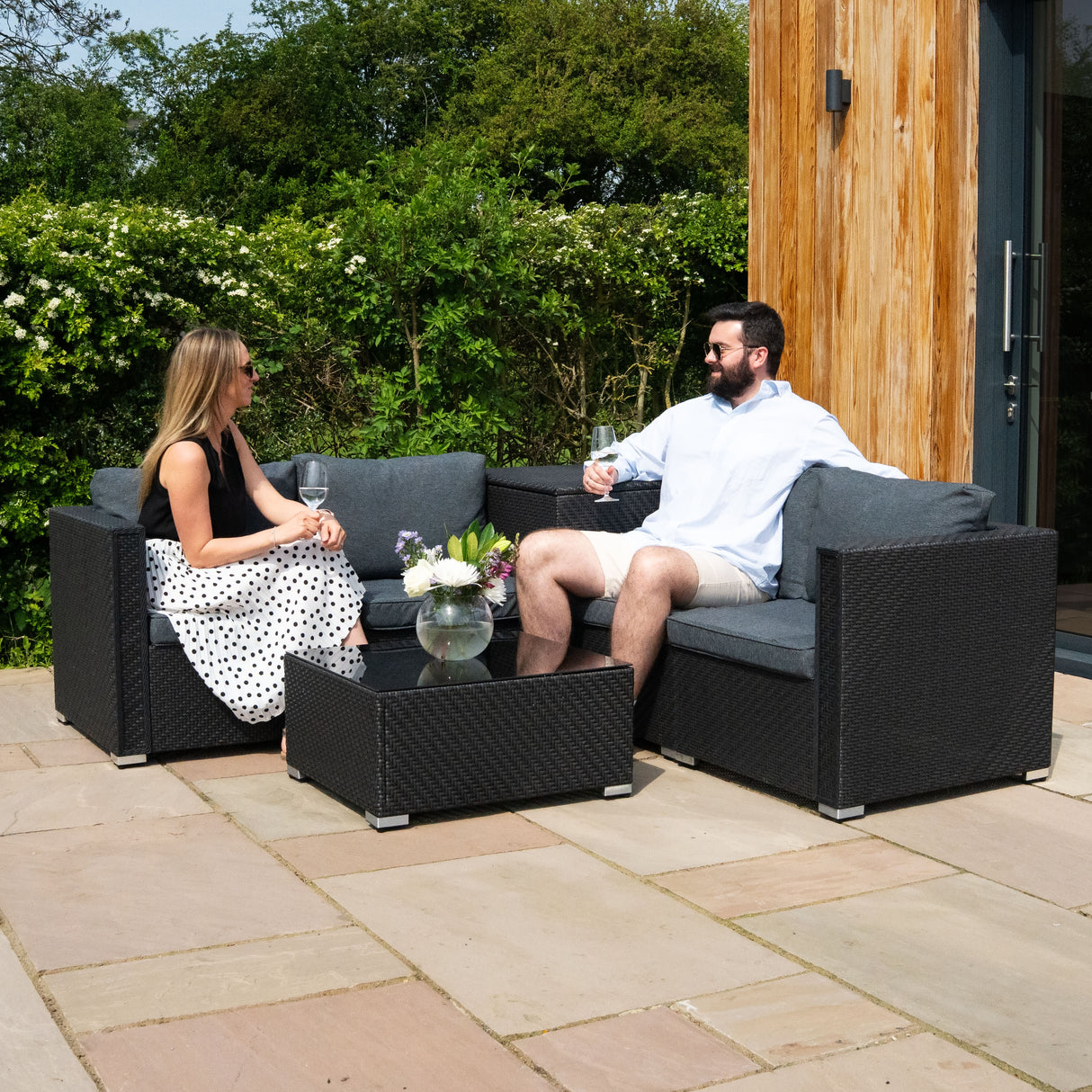 Salon de jardin modulaire en rotin noir avec coussins gris, table basse, et deux personnes profitant d'un moment convivial en extérieur