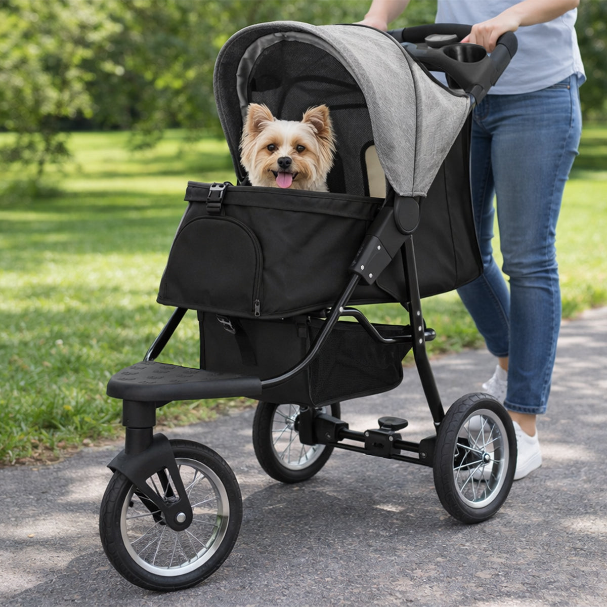 Poussette jogger pour animal de compagnie grise avec un petit chien à l'intérieur, roues noires et cadre léger sur un chemin verdoyant