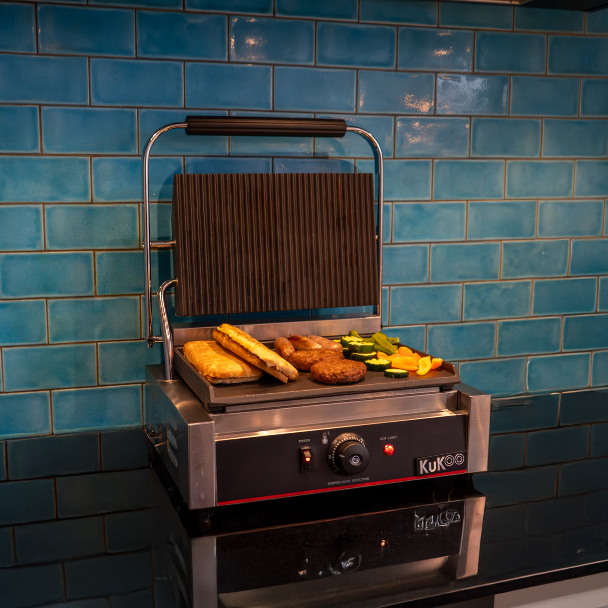 Presse à panini à plaque rainurée, finition en acier inoxydable, avec aliments grillés colorés sur une surface noire, fond bleu carrelé