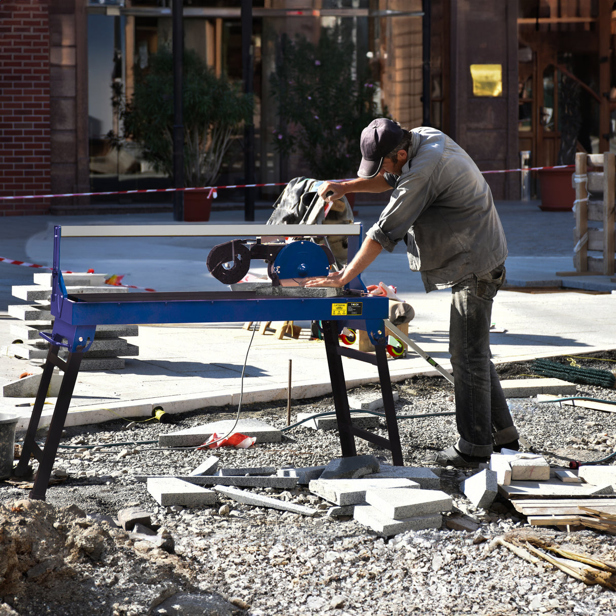 Scie de carrelage sur table bleue, utilisée par un homme en vêtements de travail, entourée de pierres et de débris de chantier