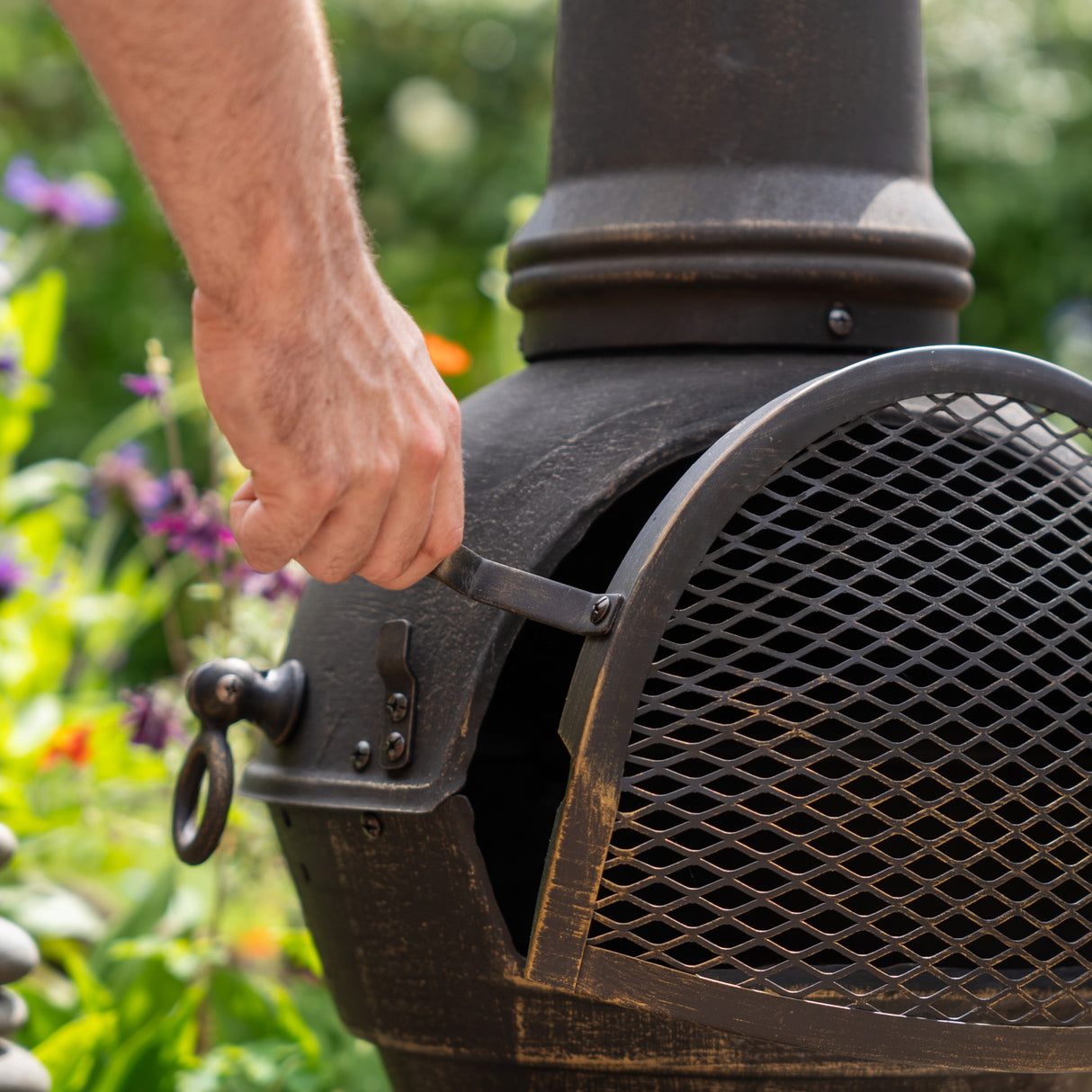 Cheminée de jardin en fonte noire avec grille en métal, main tenant la porte, entourée de fleurs colorées en arrière-plan