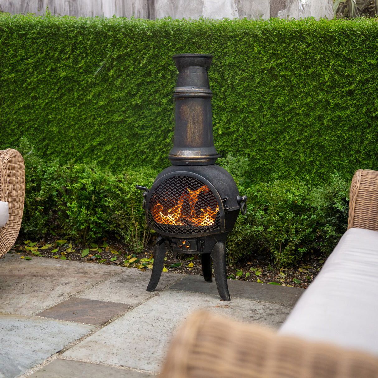 Cheminée de jardin en fonte noire, avec flamme vive, cheminée haute et grille, entourée de verdure et de mobilier en osier