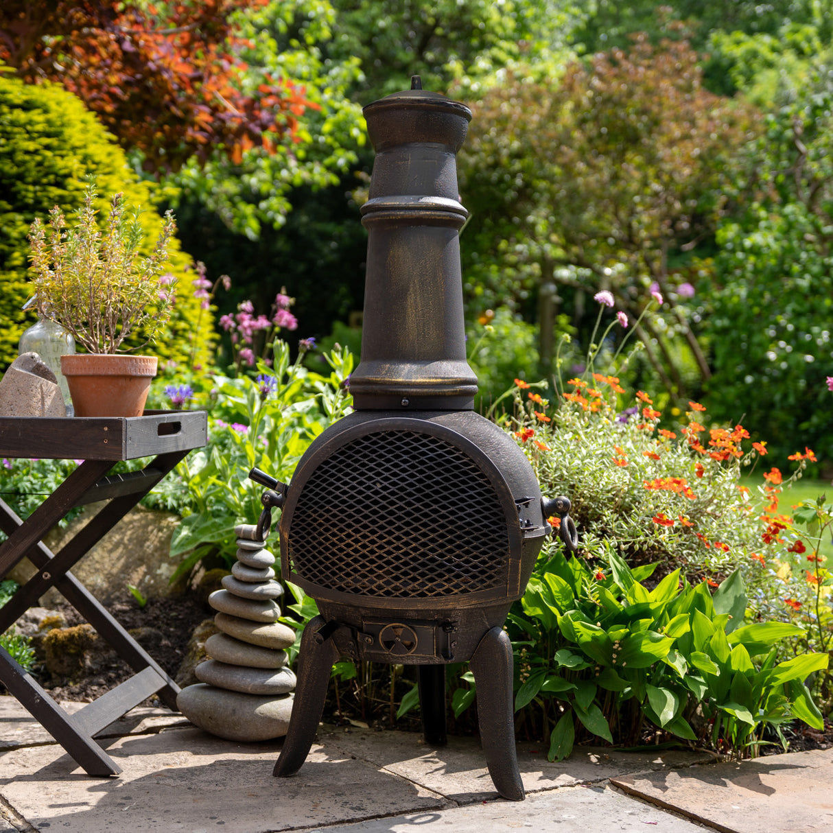 Cheminée de jardin en fonte noire avec cheminée haute, grillage décoratif, entourée de fleurs colorées et verdure luxuriante