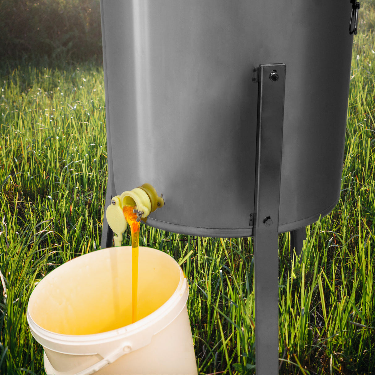 Extracteur de miel électrique en métal gris, avec du miel doré s'écoulant dans un seau blanc sur un fond de prairie verdoyante