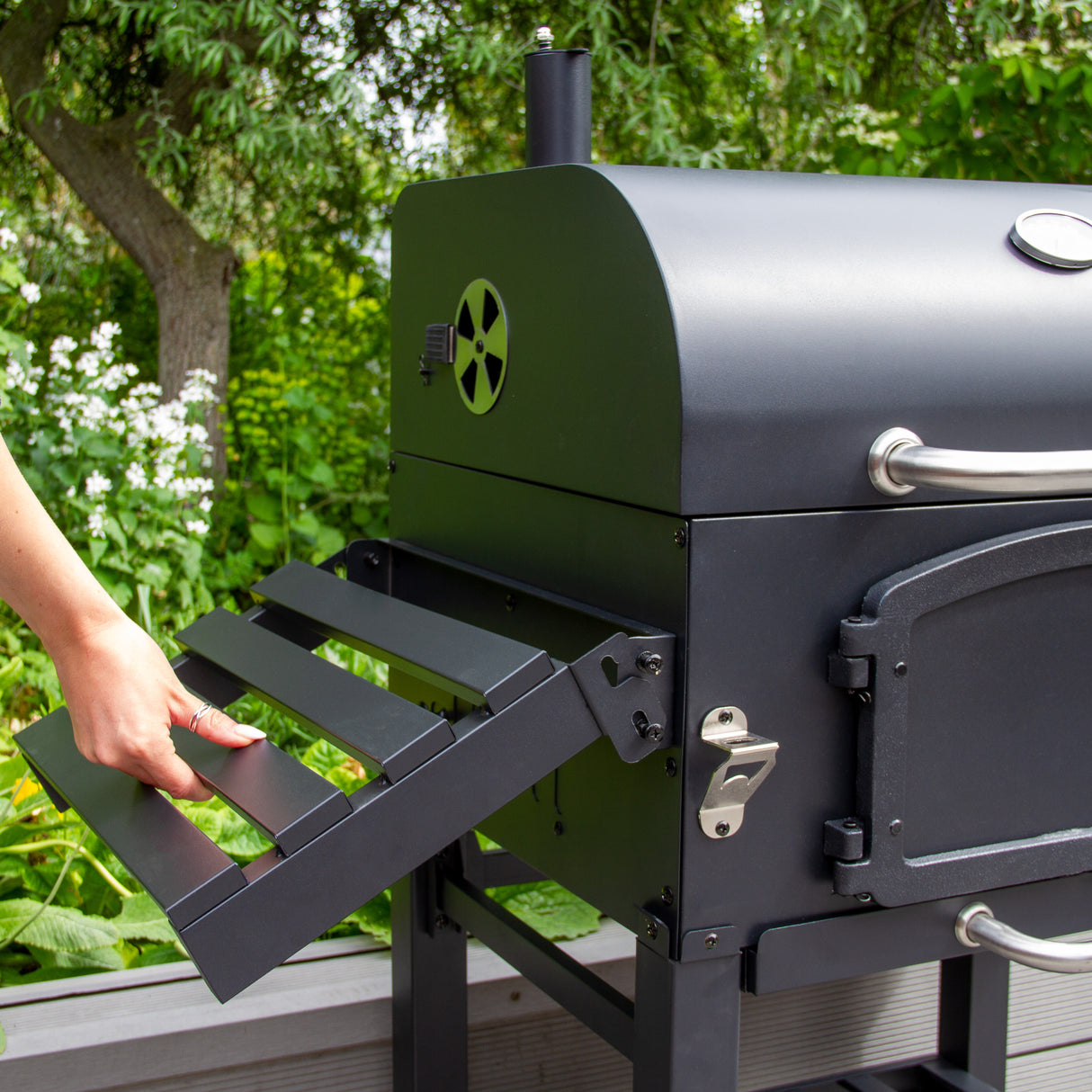Gril à barbecue et fumoir XL noir avec une grille de cuisson en métal, poignée argentée, placé dans un jardin verdoyant avec des fleurs blanches
