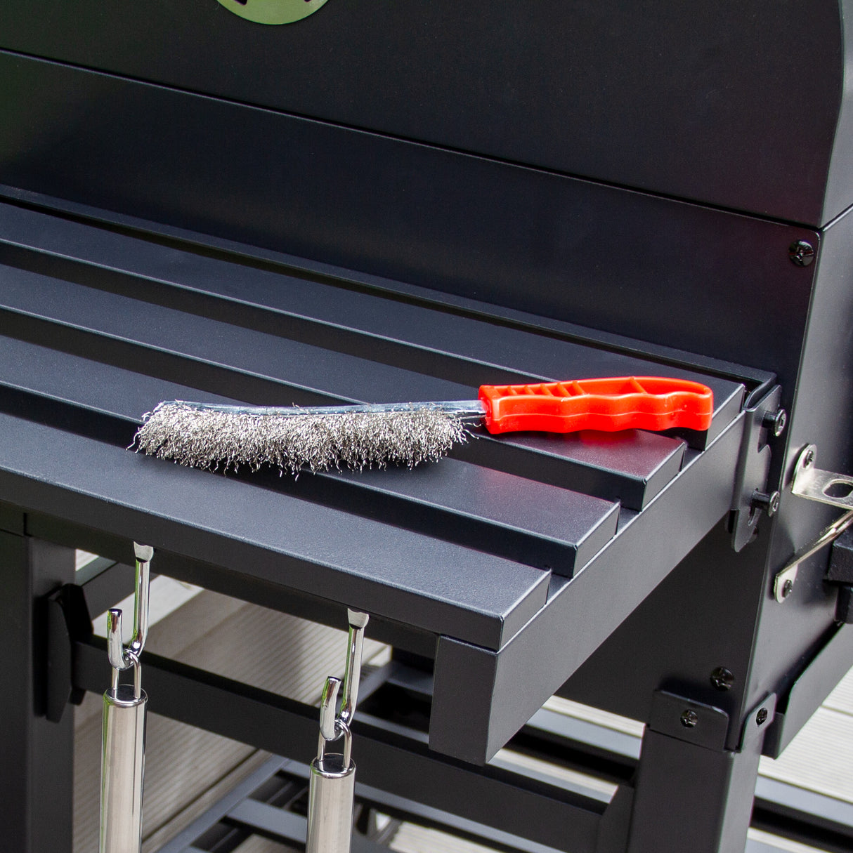 Brosse de nettoyage en métal avec poignée rouge posée sur la surface en acier noir du barbecue grill et fumoir XL