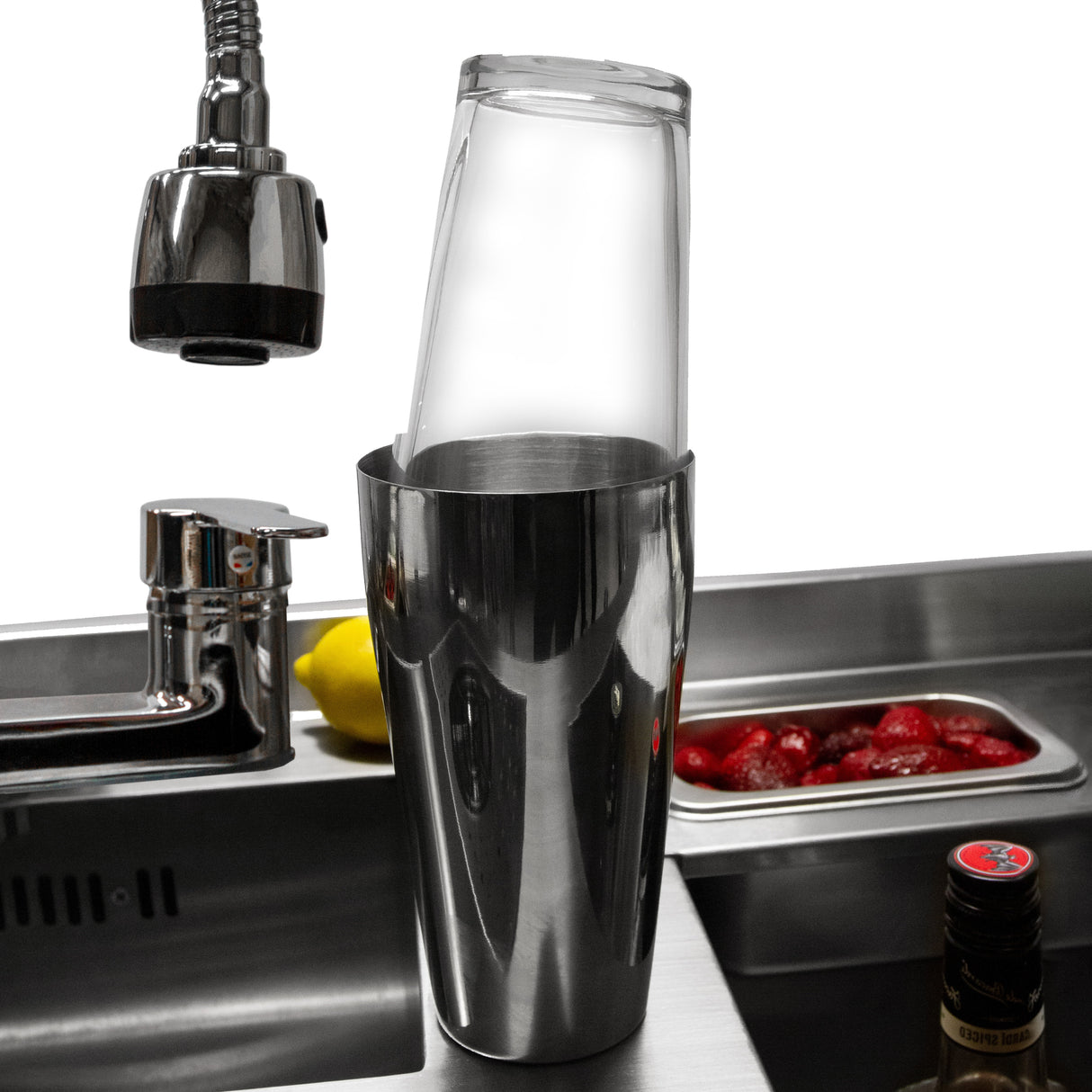 Shaker à cocktails en acier inoxydable avec un verre transparent, posé sur un bar moderne, entouré de fruits frais et d'ustensiles de bar