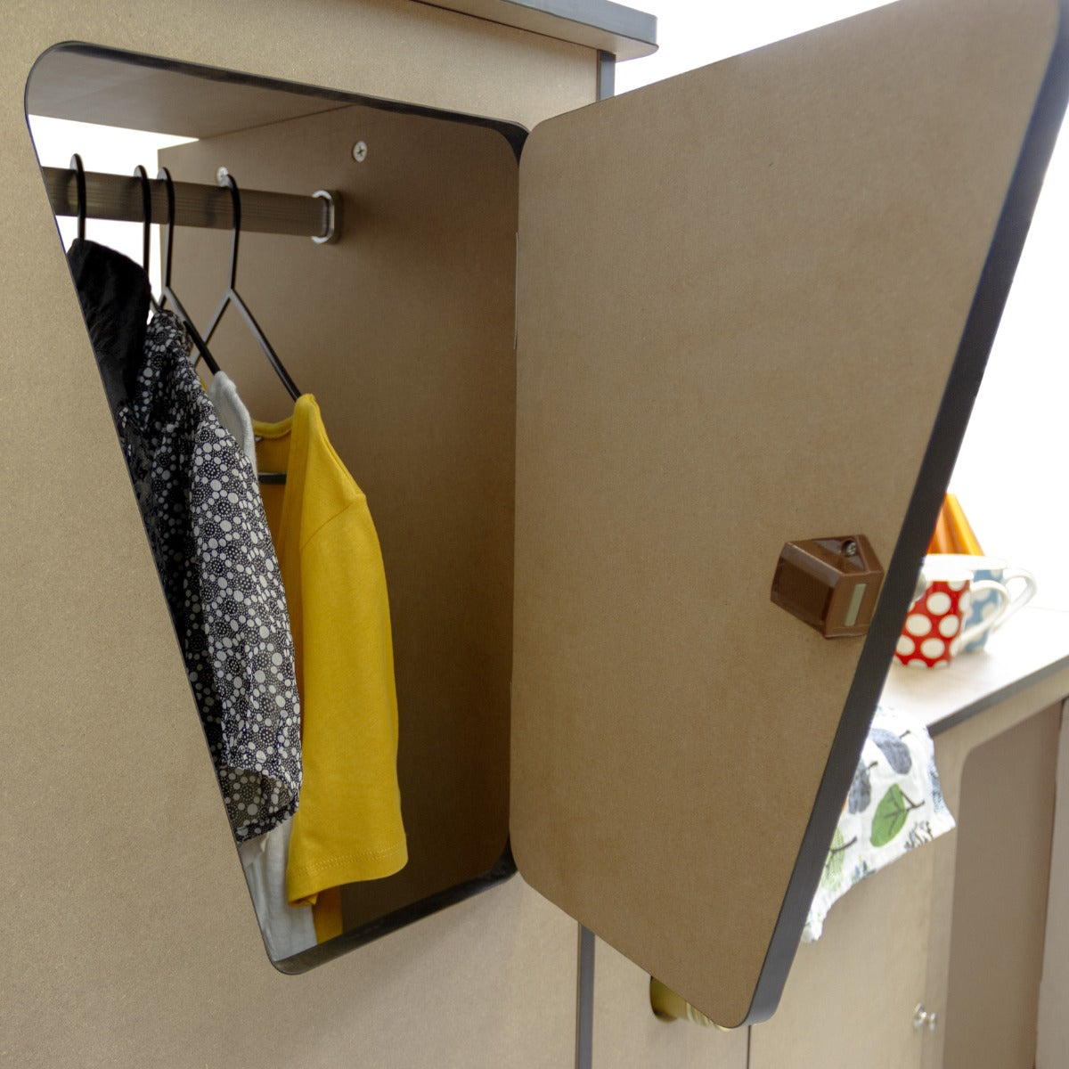 Meuble de cuisine en contreplaqué pour camping-car, avec une porte en forme de triangle, affichant des vêtements suspendus et un intérieur en bois clair