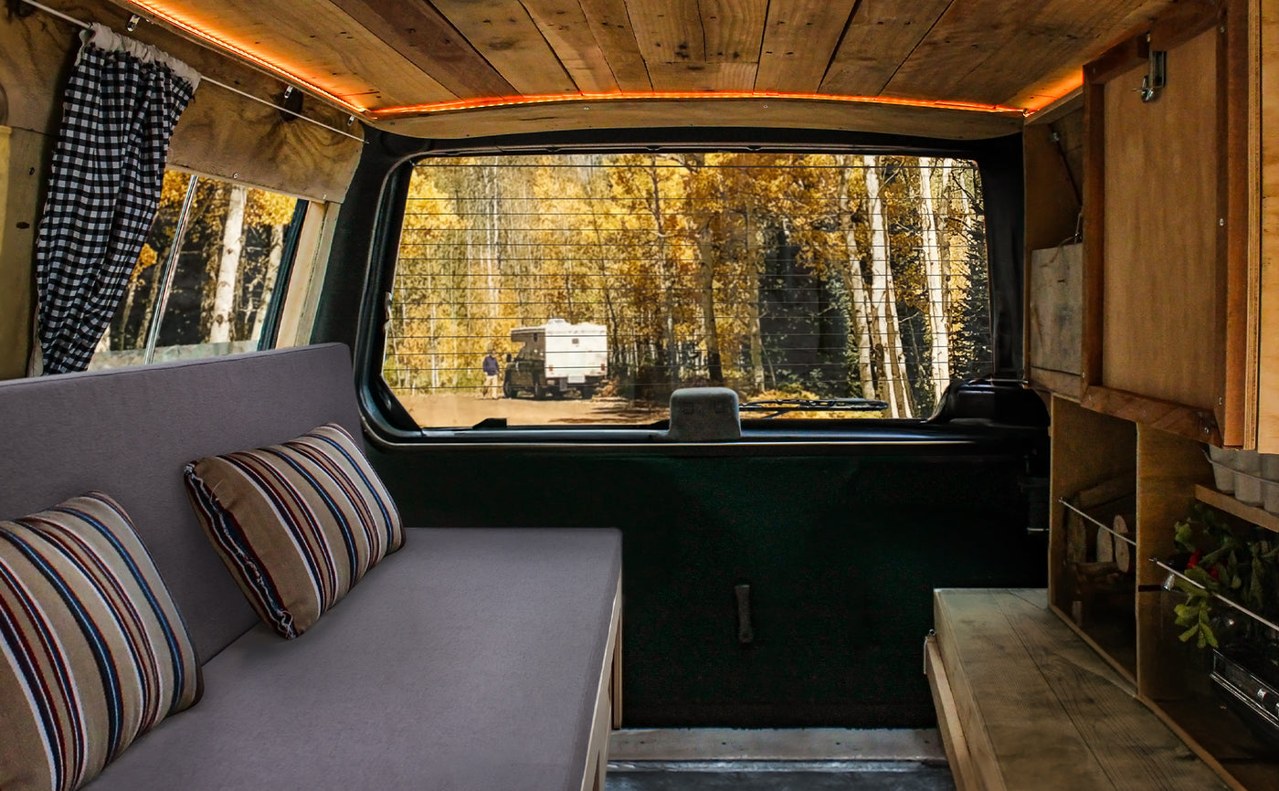 Intérieur d'un van aménagé avec un canapé gris, coussins rayés, bois chaleureux et vue sur une route bordée d'arbres dorés