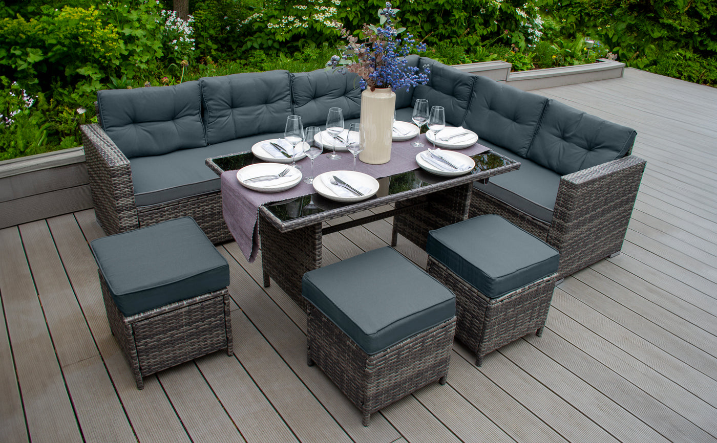 Canapé d'extérieur en rotin tressé avec coussins gris, table en verre, vaisselle élégante et décor floral sur une terrasse en bois