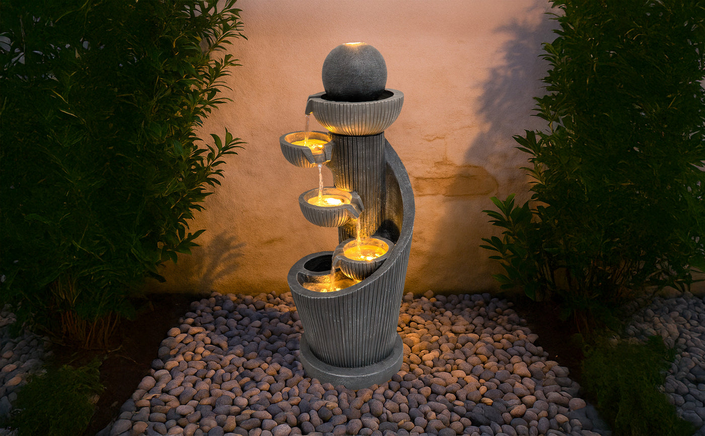 Fontaine moderne en pierre grise avec trois niveaux d'eau illuminés, entourée de galets et de verdure, créant une ambiance apaisante