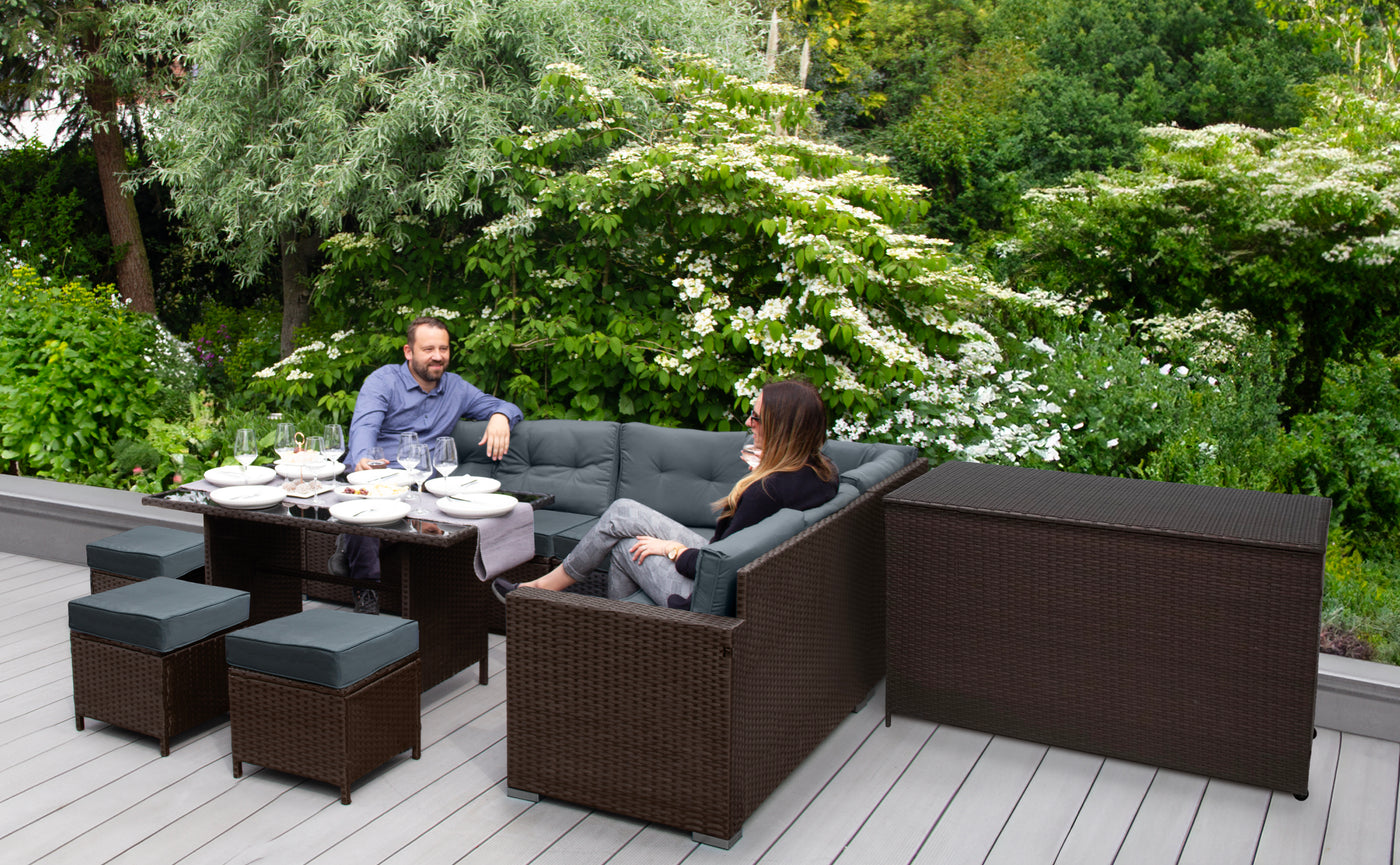 Canapé en rotin marron avec coussins gris, table basse et chaises, dans un jardin verdoyant, créant une ambiance conviviale et accueillante