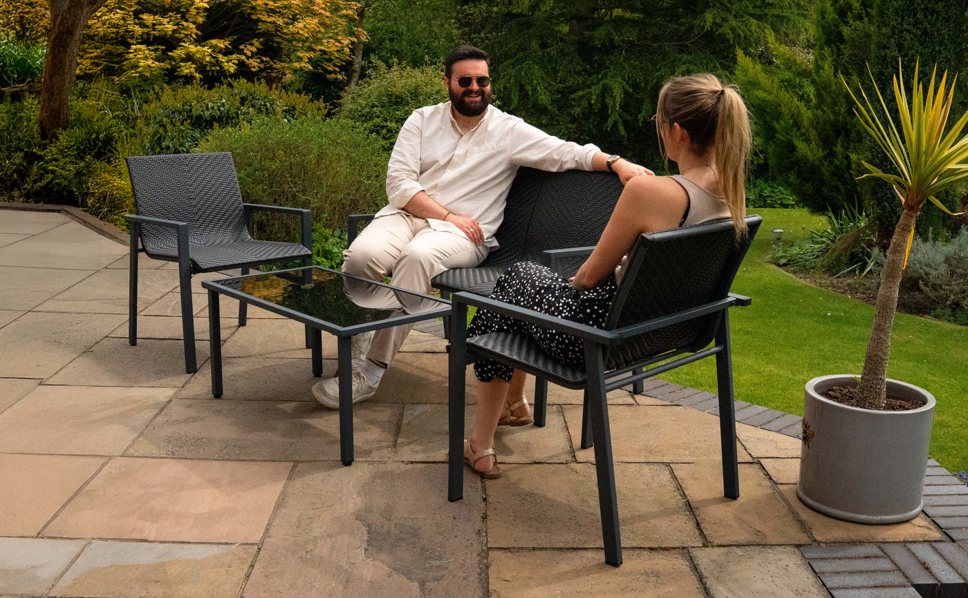 Deux personnes assises sur des chaises en plastique noir, avec une table en verre, entourées de verdure et de fleurs colorées