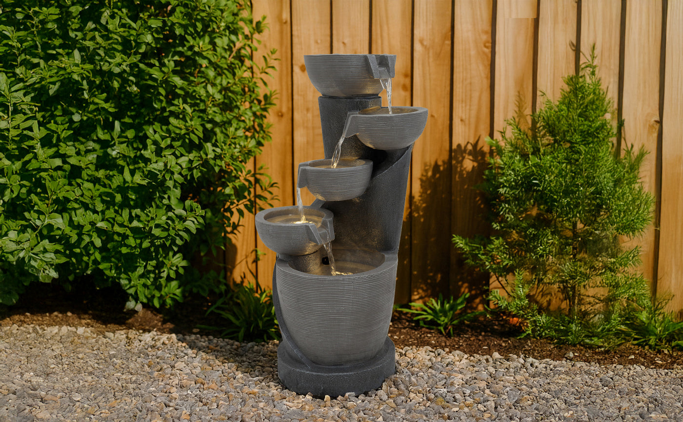 Fontaine moderne en béton gris avec plusieurs niveaux, l'eau s'écoulant doucement, entourée de verdure et de gravier décoratif