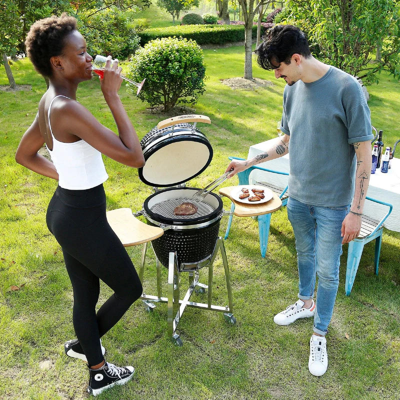 Barbecue au charbon de bois Kamado noir, 53,3 cm, avec des steaks grillés, utilisé par un homme et une femme dans un jardin verdoyant