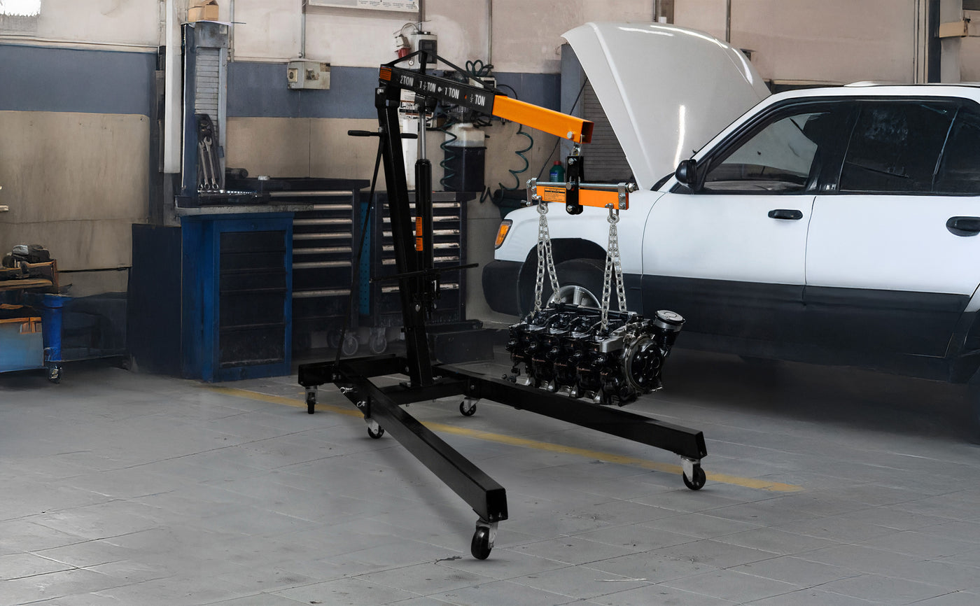 Grue de moteur noire avec chaînes, utilisée pour soulever un moteur dans un garage, avec un véhicule blanc en arrière-plan