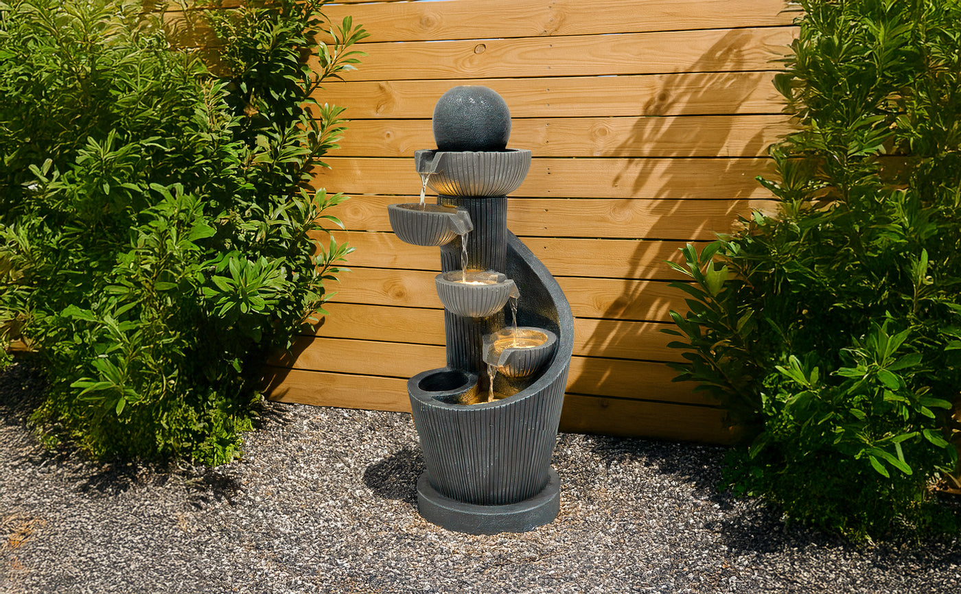 Fontaine moderne en pierre grise avec plusieurs niveaux, eau s'écoulant doucement, entourée de verdure et de gravier décoratif
