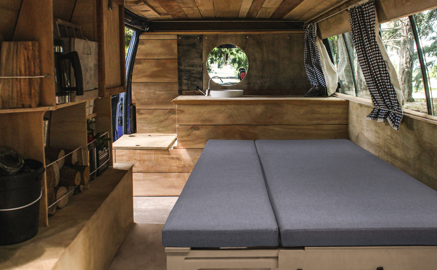 Intérieur d'un van aménagé avec un matelas gris sur une base en bois, fenêtres avec rideaux à carreaux et espace de rangement en bois naturel